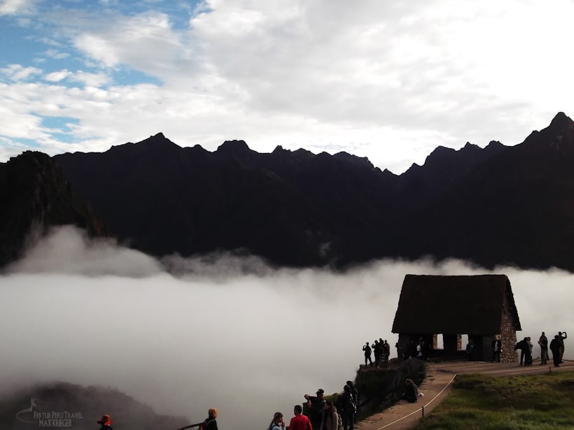 Casa del Guardián de Machu Picchu envuelta en niebla matutina con siluetas de turistas y cordillera andina al amanecer, atmósfera mística en ciudad perdida de los incas