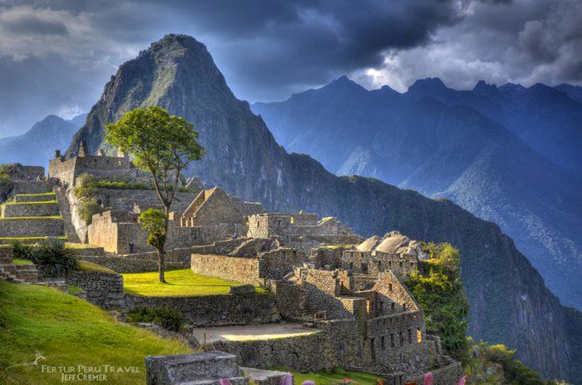 Plaza Pisonay de Machu Picchu con iluminación dorada y cielo dramático, montaña Huayna Picchu al fondo, terrazas verdes y arquitectura inca, fotografía de Jeff Cremer