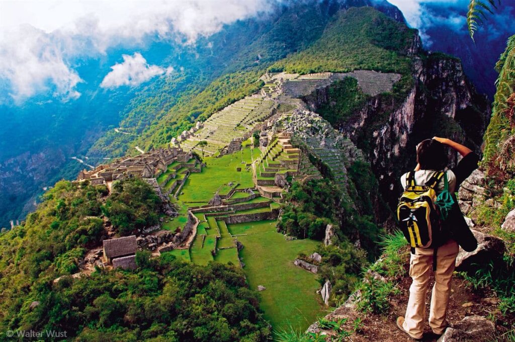 Fotógrafo contemplando vista panorámica de Machu Picchu desde Huchuy Picchu con mochila amarilla, terrazas verdes y montañas andinas cubiertas de vegetación, fotografía de Walter Wust